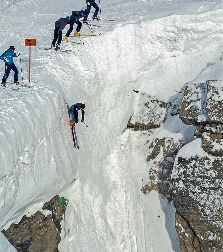 an image of a skier vertical dropping into to a famous couloir (Corbets in Jackson Hole) with skiers teetering on edge looking over the precipice looking at skier. Drop in is huge, metres upon metres, you can't see the bottom where skier would land