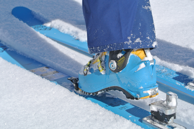 blue skis, blue boot, blue trouser cuff clipping into a touring binding on a few mm of fresh snow. Ad for bindings