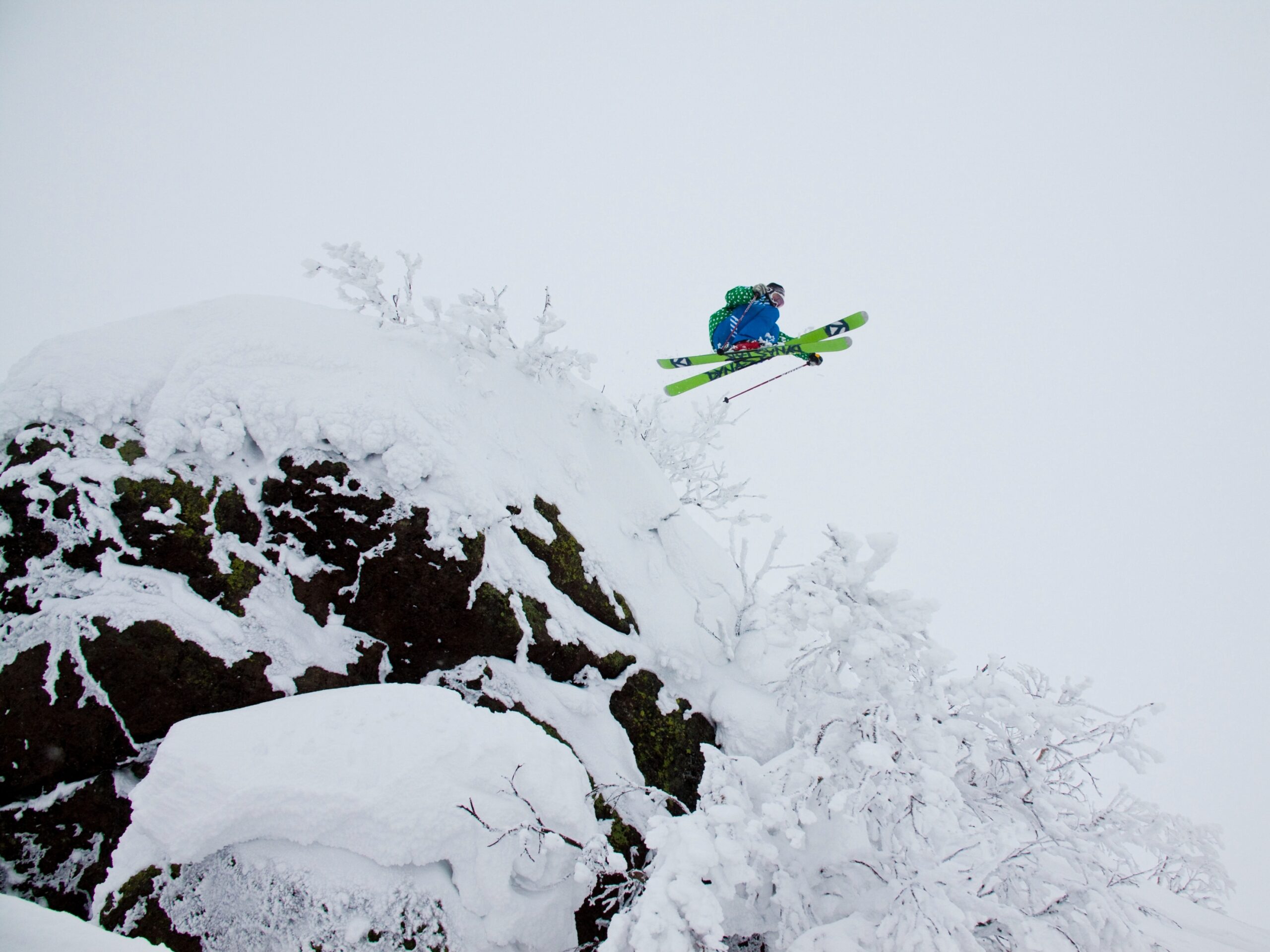 green base of skis is the only real colour in this white-room shot of a skier (in dull blue) take a huge huck off a rock, with the photo taken from beneath