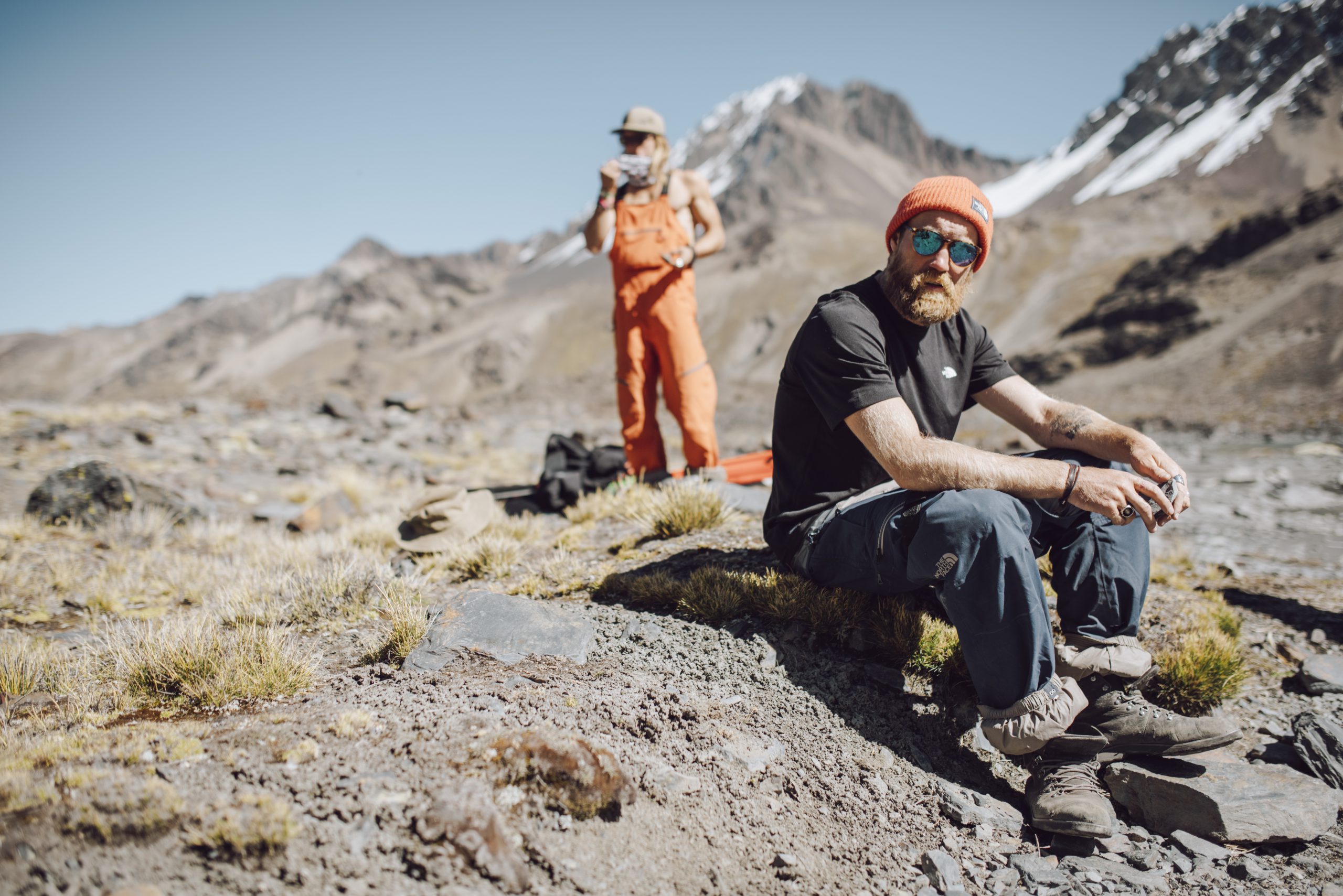 sam smoothy sits on a rick at high altitude in a t shirt, with someone else standing in background. It looks hot