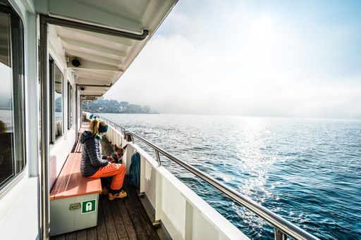 a woman wrapped up warm sits on the benches of a ferry boat outside, watching over blue water as the sun shines down