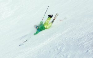 A skier in yellow and orange has fallen and is sliding down a steep hill, equipment akimbo