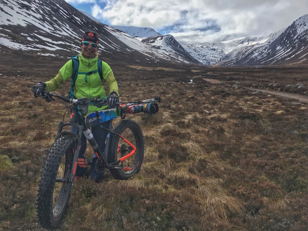 a fat biker stands on grassy land, surrounded by snow-streaked mountains, holding the handlebars of a bat bike packed with skis, boots clipped in.