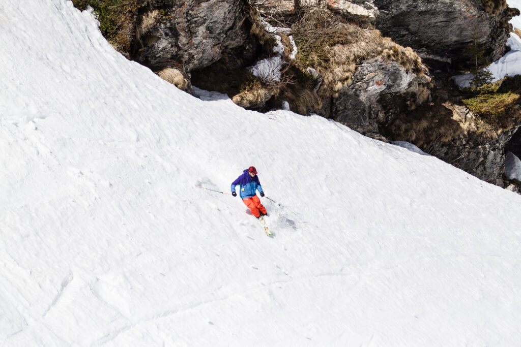skier in orange pants and blue jacket skis on sketchy crud snow in front of rocks