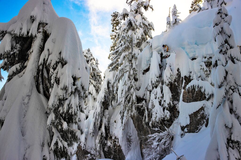 skier hucking off a serious cliff drop, tonnes of snow on laden trees