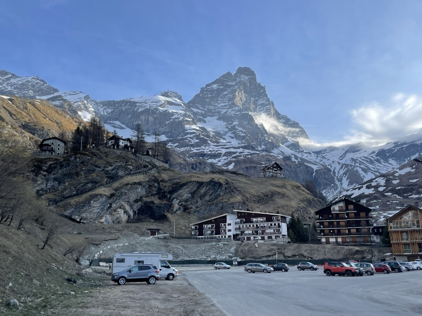 Cars and vans are parked in a carpark at the foot of the Matterhorn/Monte Cervino at first light. Traditional wooden chalets are seen and the mountains are streaked with snow (mostly melted)