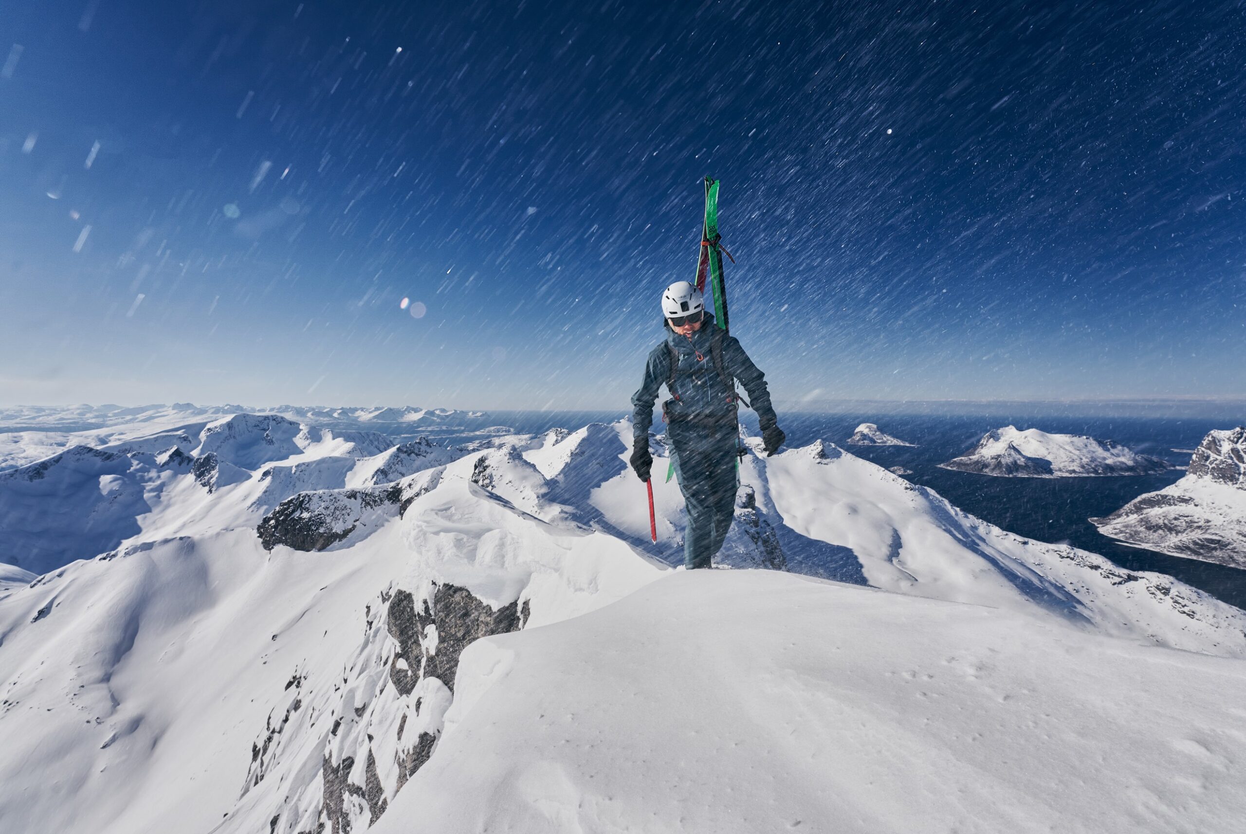 man geared up with skis straped to back reaches summit of mountain on foot. looks like norway with sea and mountains rising out of water, with small mountain islands. snow is coming down out of a blue sky