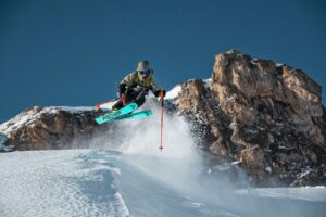 skier takes serious air on a turn against a backdrop of rock and snow spray. you can see the turquoise underside of his skis and orange poles