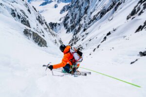 skier sam smoothy in orange jacket skis down a deep couloir in very snowy environment so steep he's roped up, visible in foreground of shot