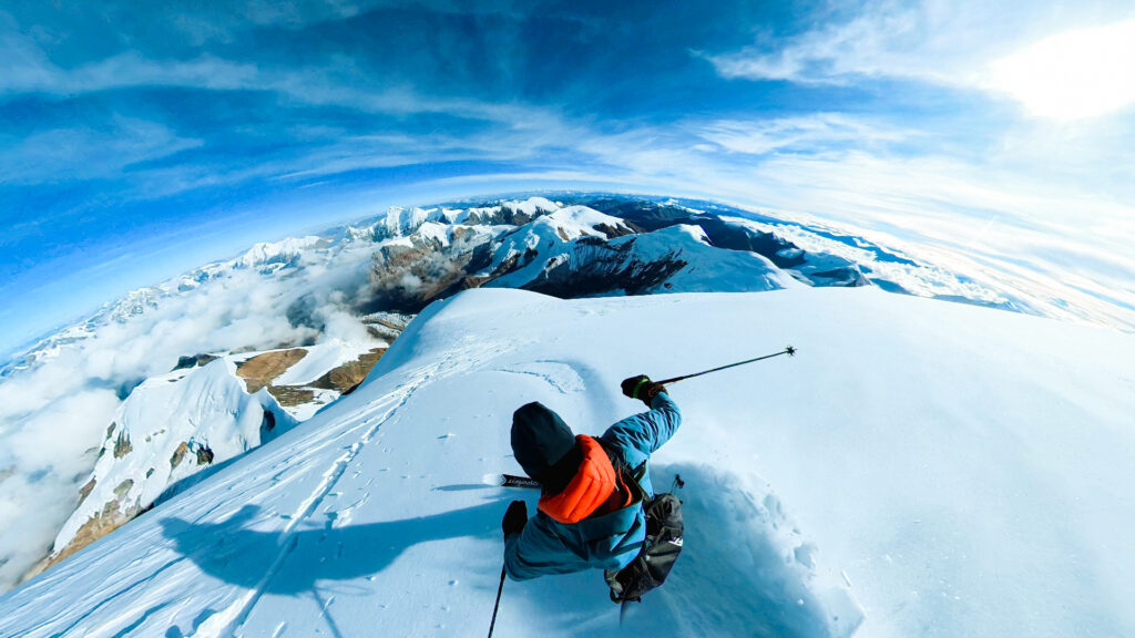 a skier makes deep tracks in snow at the top of a mountain, surrounded by lower peaks also covered in snow. image taken from above and behind the skier