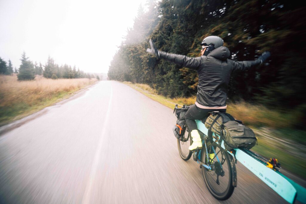 skier on a bike cycles, geared-up, skis attached lengthways with bike between skier's legs, as he rides hands-free along a flat, pine-tree lined road