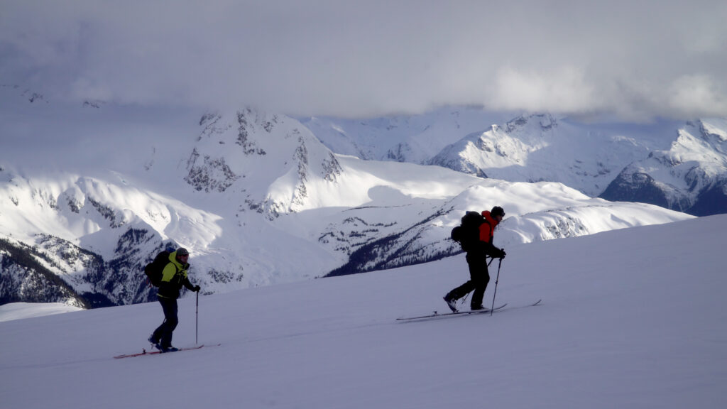two skiers skin up a gentle hill but high, with mountain tops in the background.