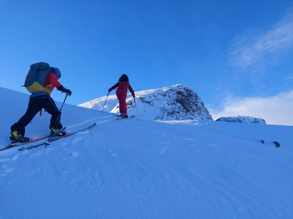 two ski tourers climb snowy mountain in the shadow, nearing mountain top, against a blue sky