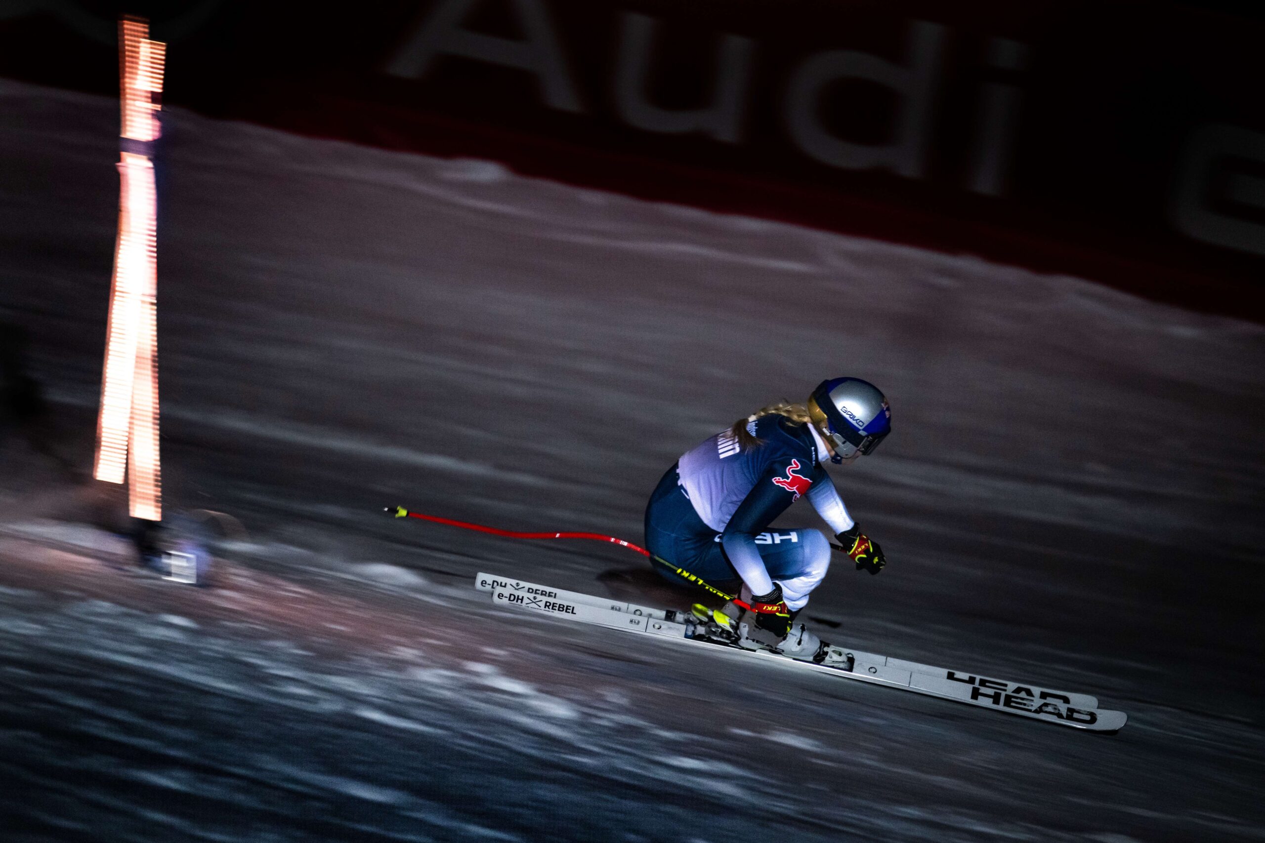 retired ski racer lindsey vonn on downhill skis wears red bull branded race gear on a course in kitzbuhel. it's dark, at night, and the skier is tucked, making a turn in air