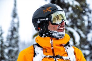 sam kuch ski athlete head shot wearing helmet and goggles with beard covered in snow with trees behind out of focus