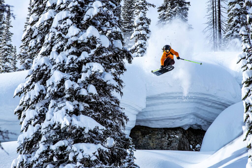 skier sam kuch jumps with rail grab off a mushrroom drop rock, with snow so thick you can see layers towards fresh snow and tree laden with snow