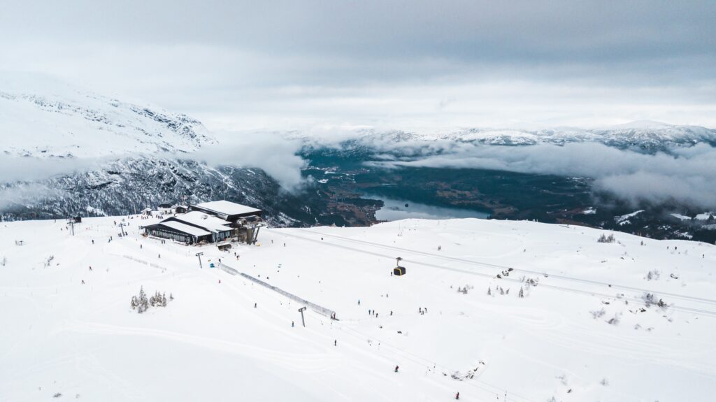 a zoomed out view of Voss ski resort in norway with blue coloured mountain strip sandwiched between white sky and snow. a cable from a ski lift is in sight with skiers dotted around, as well as on a drag lift.