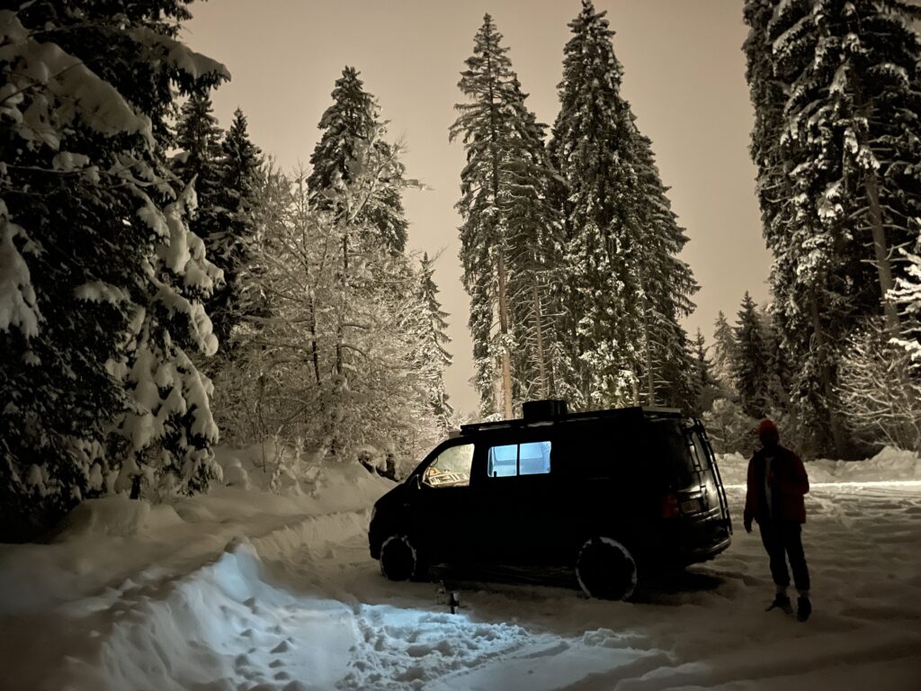 van in snowy evnironment shot at night with tall snow-laden trees surrounding. a figure stands next to the black campervan as a silhouette