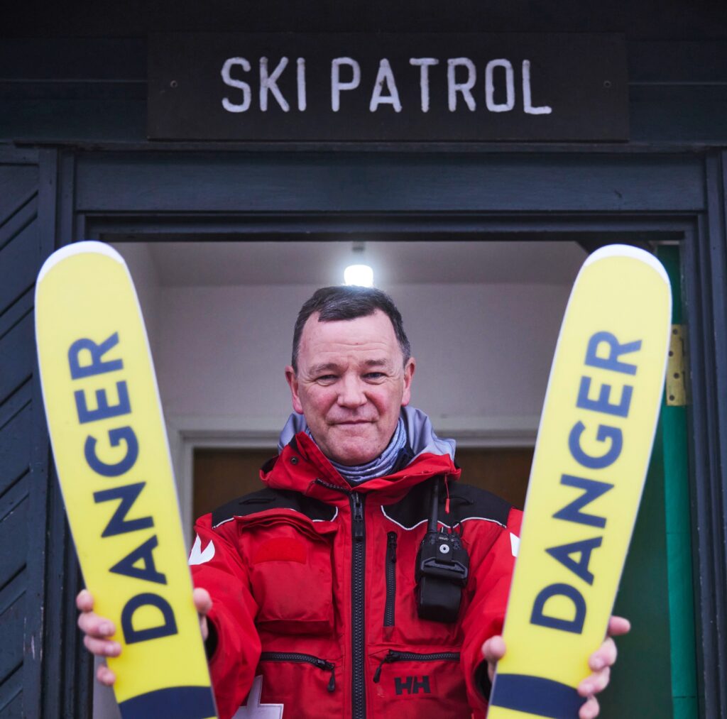 patroller in red jacket stand sunderneath sign holding skis of which the bases read 'danger' in black on yellow