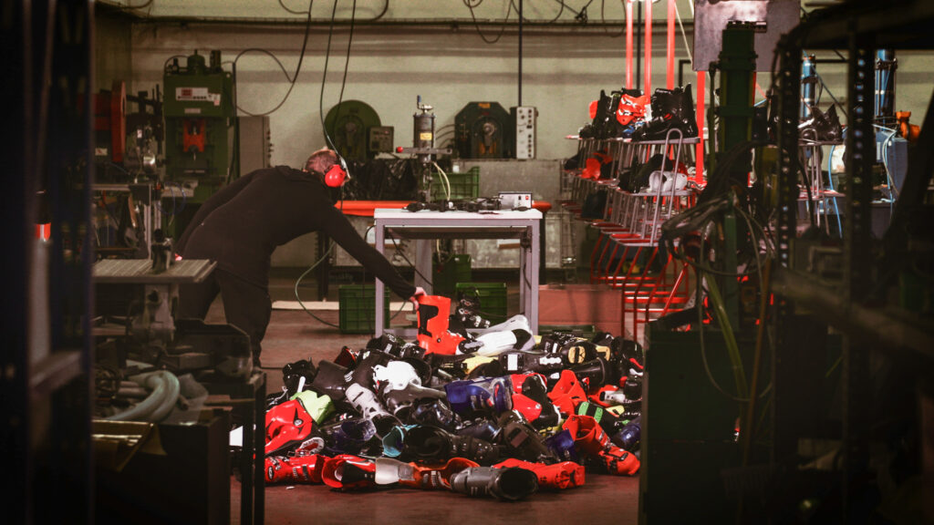 pile of ski boot shells lie in the middle of a factory floor, multi-coloured (mainly red and black) with a man wearing ear defenders placing a red boot shell to the top of the pile. The rest of the room containd machinery and shelves of more ski boot shells