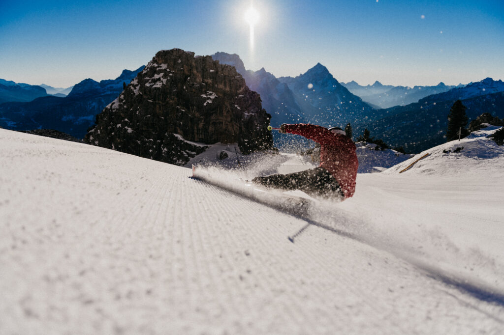 a skier makes a right turn carve spitting up dust and spray which the camera captures, positioned close to the groomed piste. In the distance a dolimitic limestone tower, splitting the piste, is free of snow on a sunny day