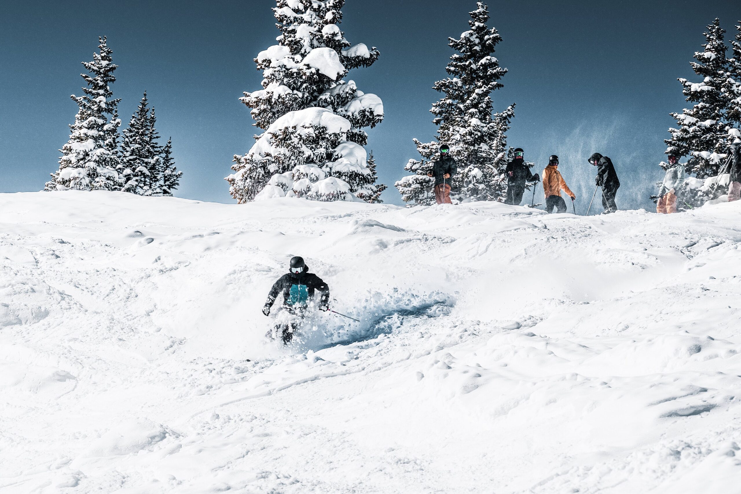 a skier on choppy, heavy looking snow watched over by a handful more skiers at the top of the slope