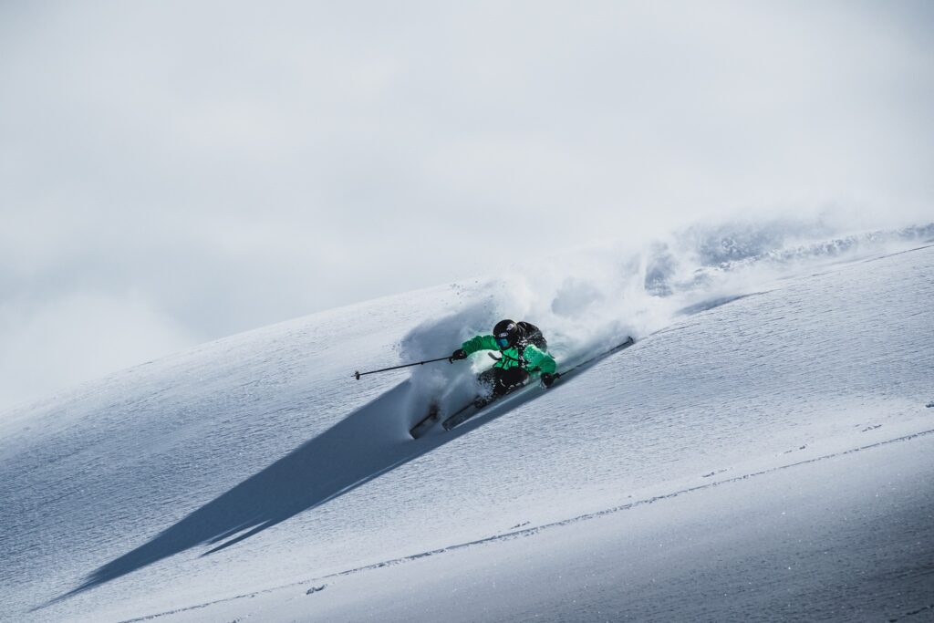 skier in green on fresh snow, spraying on a left turn at what looks to be the top of a mountain, off-piste, with a white cloudy background