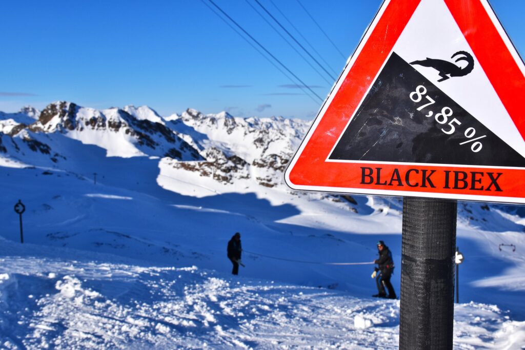 warning ski sign with steep slope 87% slope is shown on a triangle sign with ibex leaping printed on, in front of a ski area with two skiers visible in background