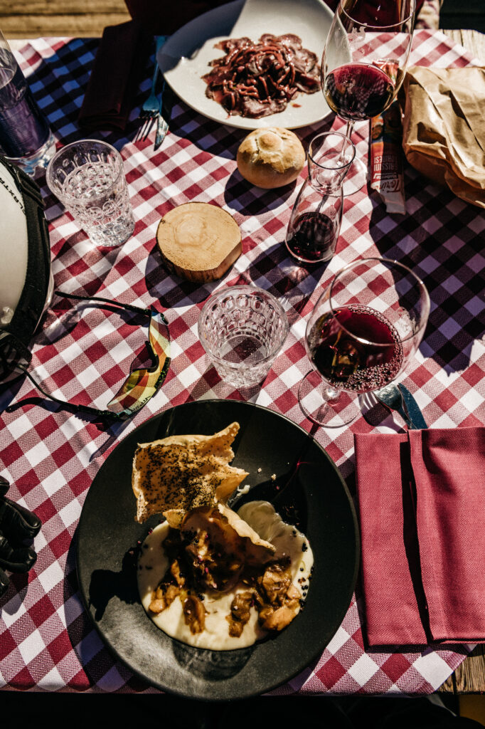 a plaid tablecloth with red wine, bread rolls, sunglasses and plates full with local Italian Ladin mountain food