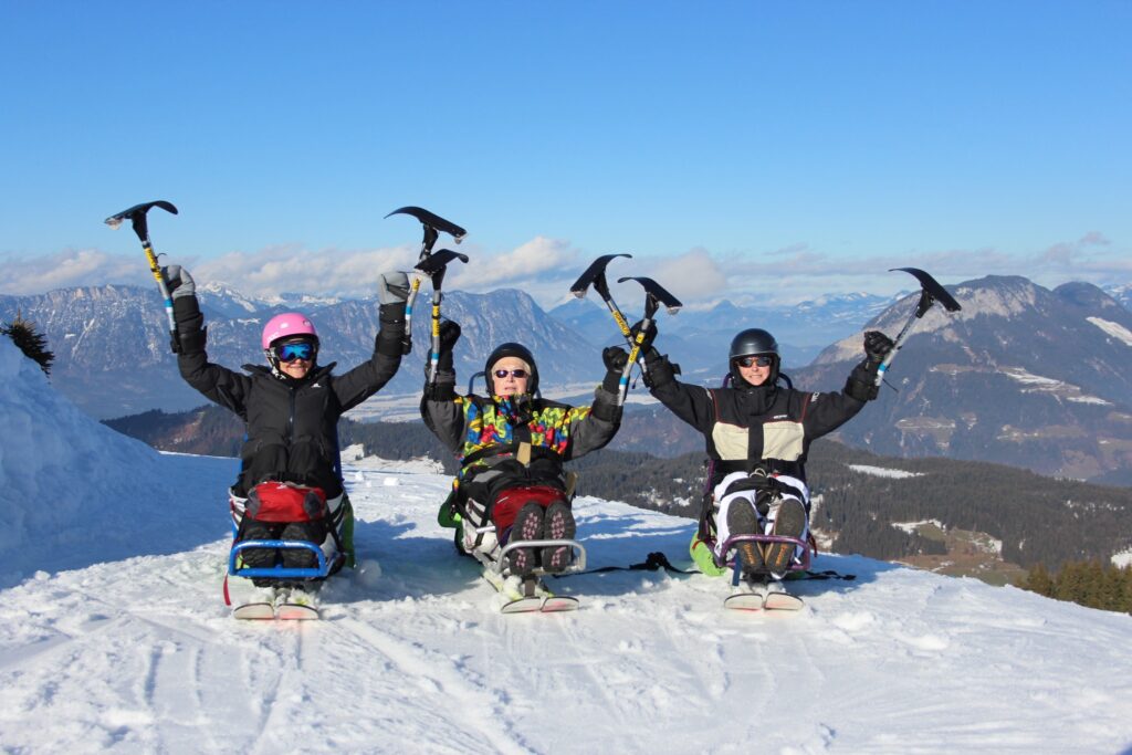 three sit skiers are on the top of a mountain with a wide valley below in the distance, with adaptive skiers' arms (and hand skis-blades in air)