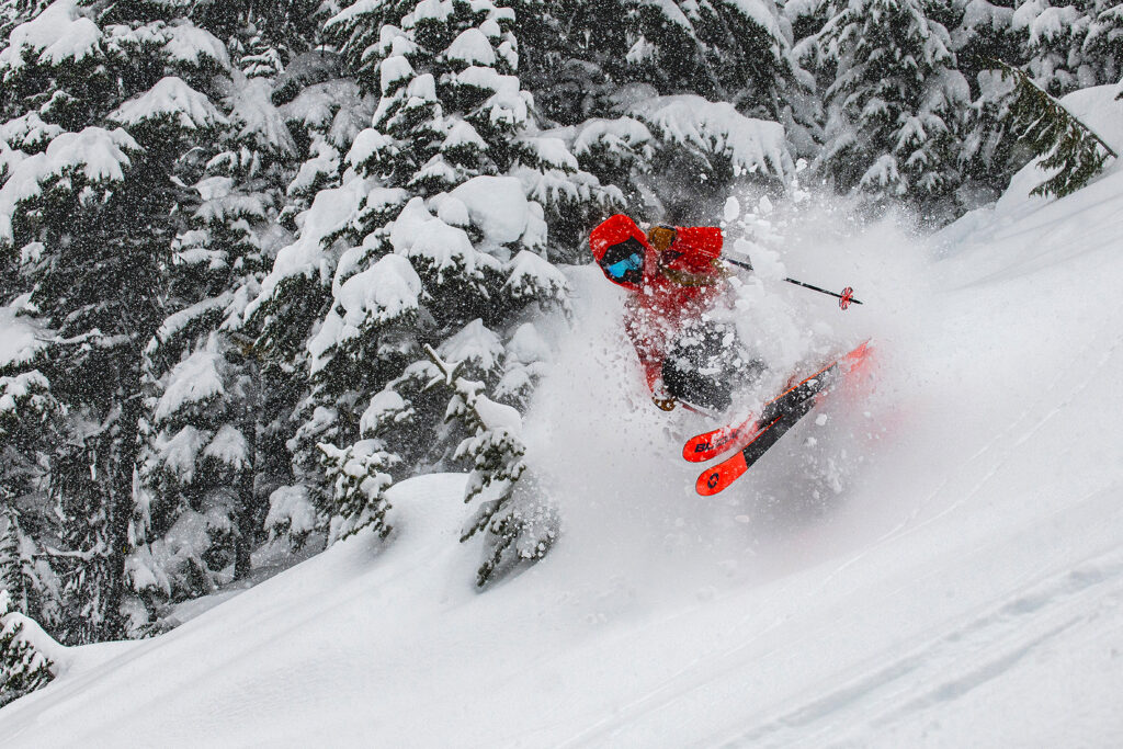 skier in red, with red on ski base tips and tails, jumps over little (invisible) bump through snow-laden trees creating a powder cloud