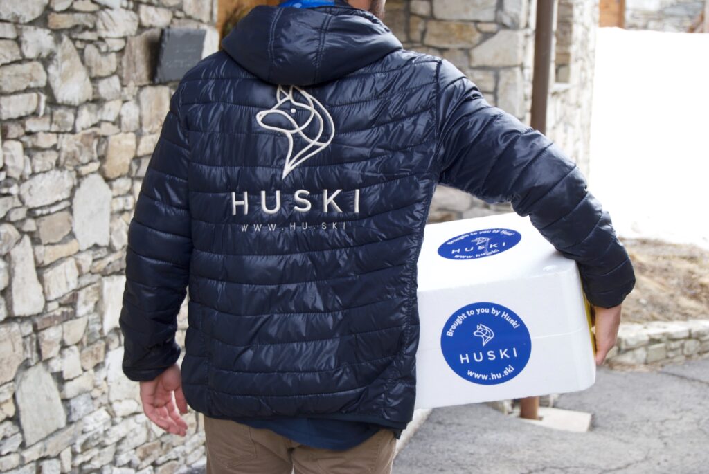 man in puffa jacket delivers food from Huski delivery service, carrying box to a stone chalet