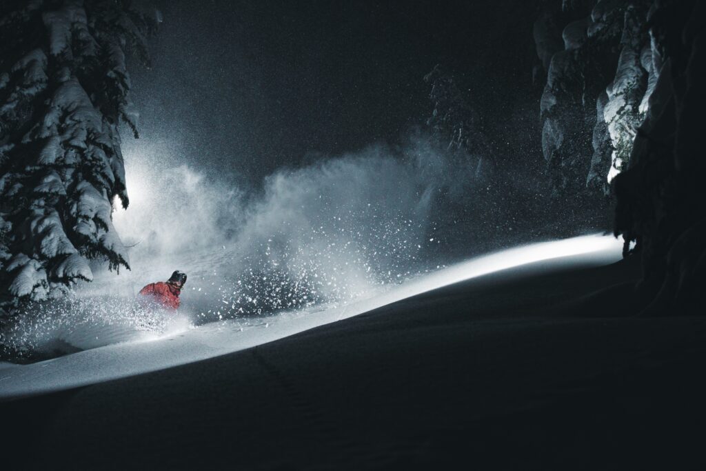 skier or snowboarder (can't tell which from snow spray) in red jacket skis deep powder at night with an interesting light cast across their line. Two trees frame the picture laden with snow