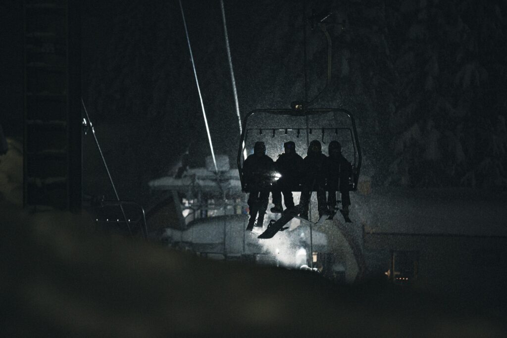 four skiers and snowboarders sit on a chairlift at night, lit up by a light at base station