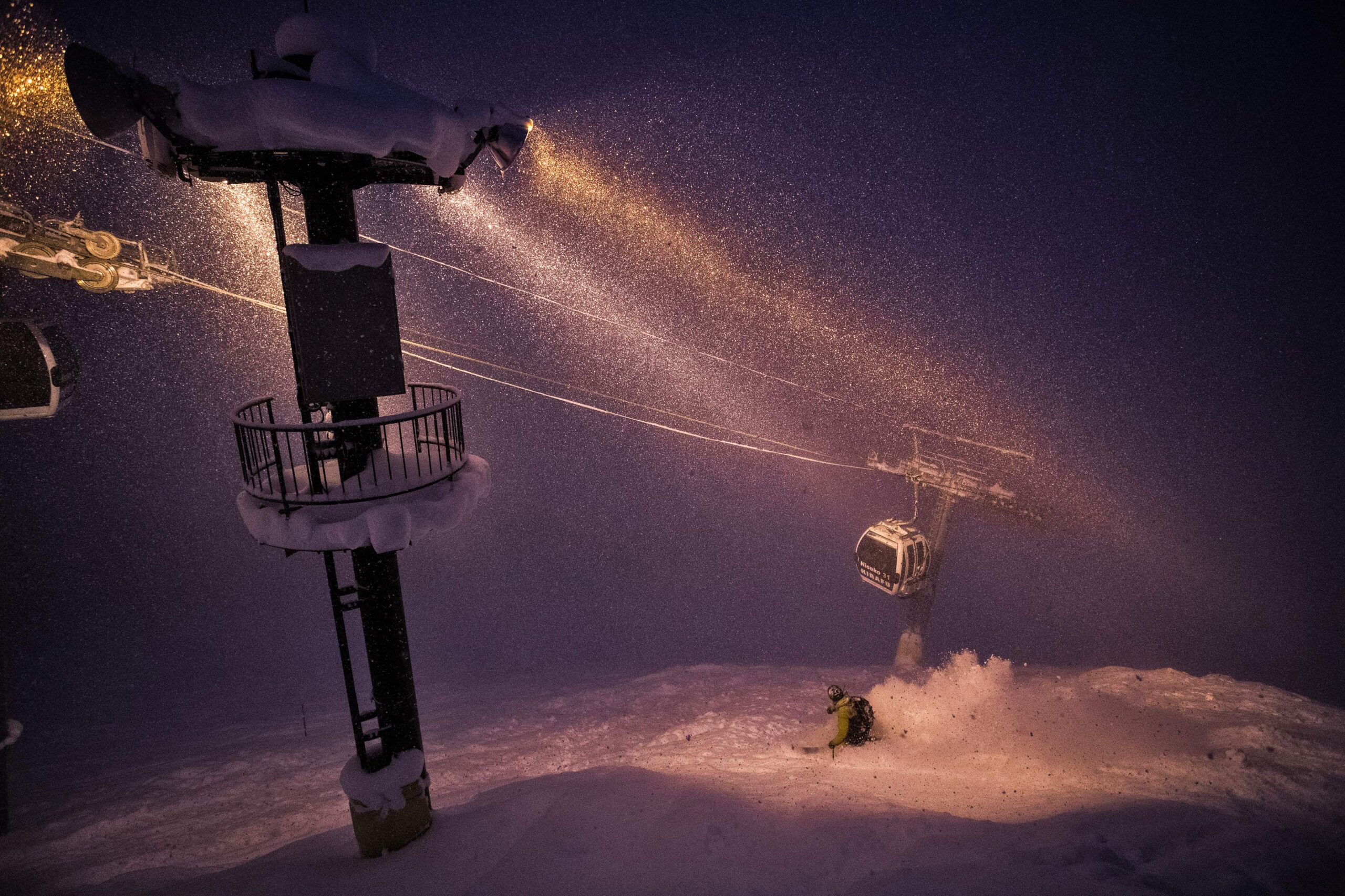 a gondola pylon shines light on wires and gondola beneath, but also on skier in green, night skiing off piste, under the lift line
