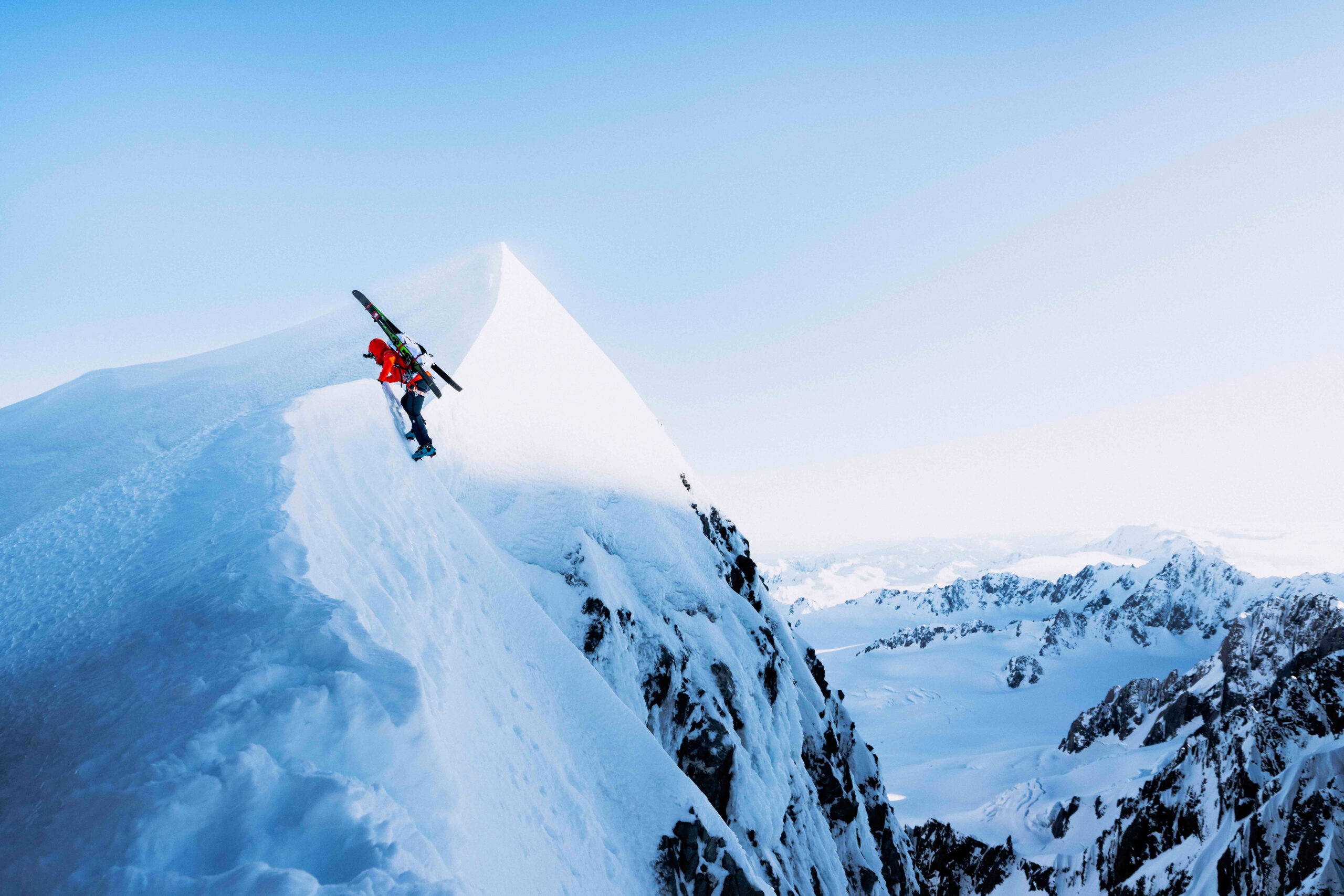 skis strapped to back, reaches apex of a snowy peak on a blue sky day by climbing