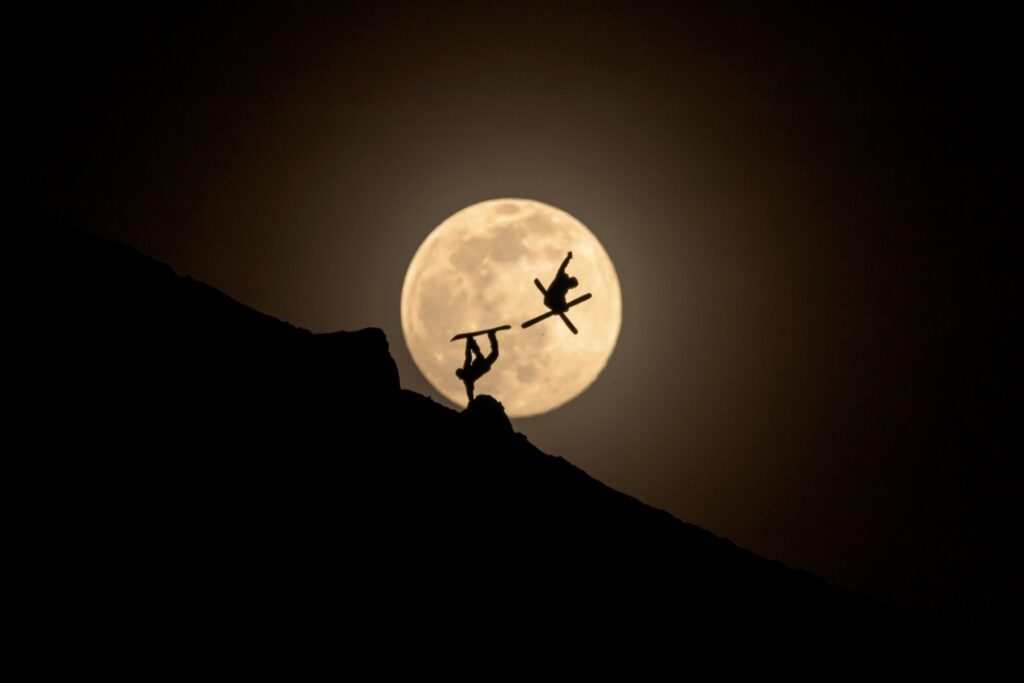 skier and snowboarder perform tricks in front of the moon, visible as silhouettes against the huge, low hanging moon