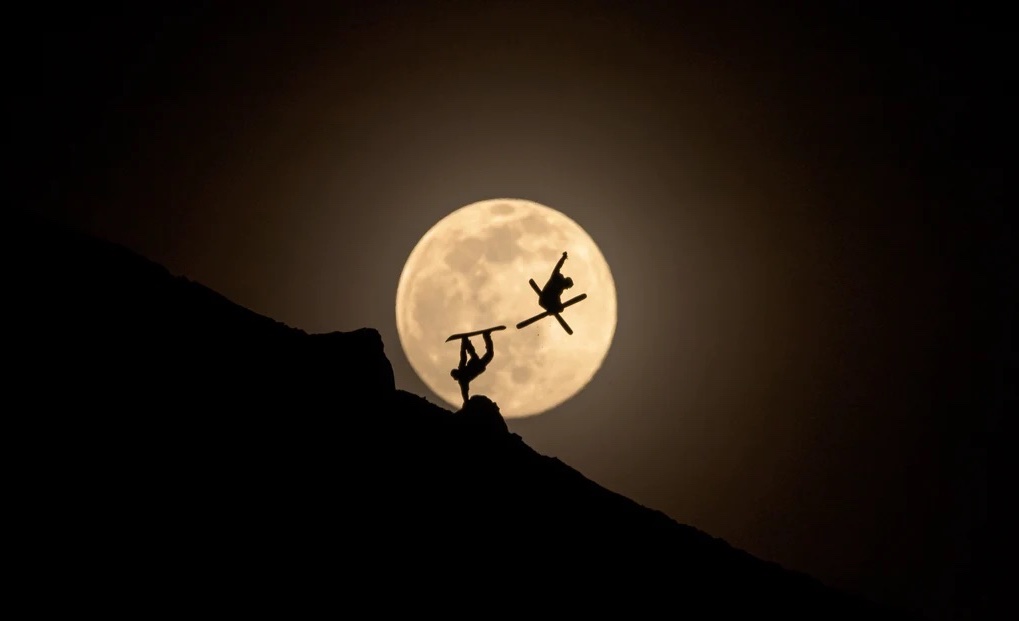 skier and snowboarder perform tricks in front of the moon, visible as silhouettes against the huge, low hanging moon