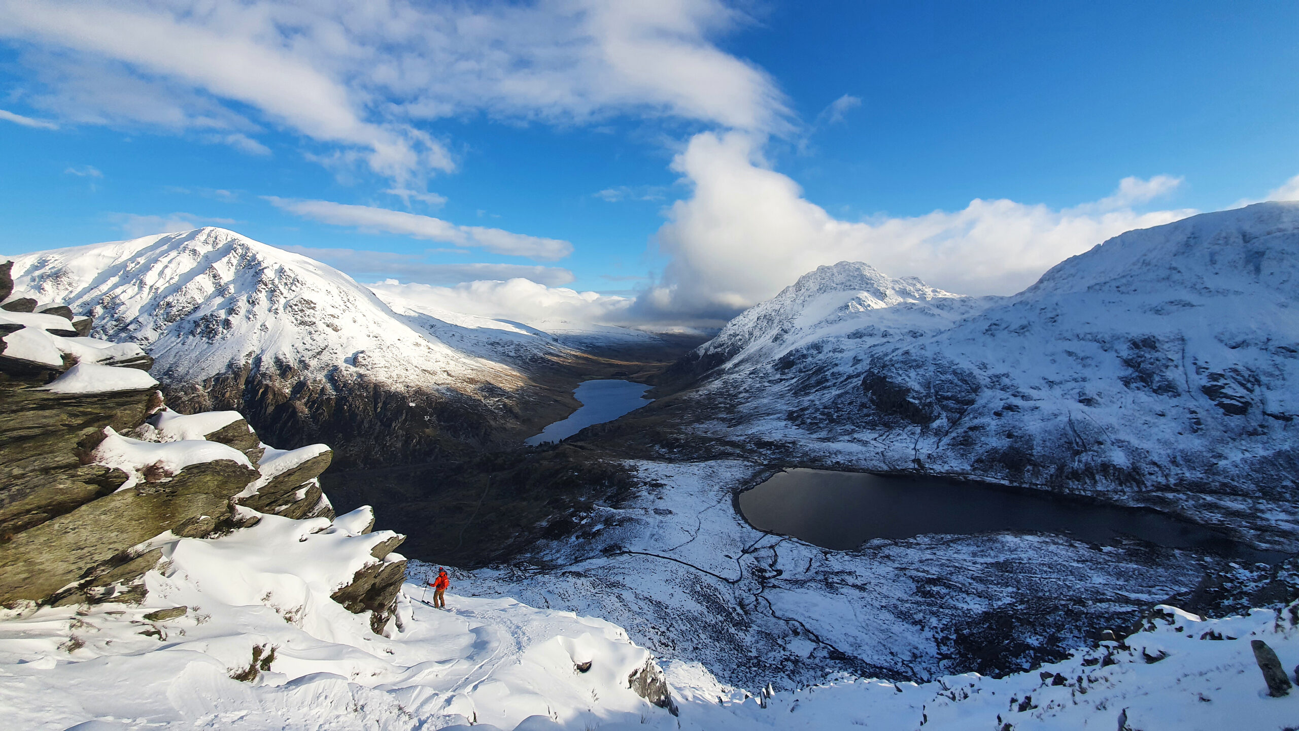 welsh landscape covered mostly in snow with a lake down in valley in far distance, with a skier in red standing under a cliffy rock in the nearer-ground