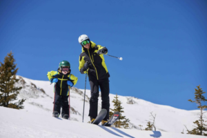 father and son on slopes together on skis as dad points out something to small son with pole