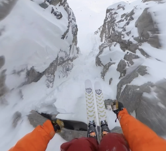 black crow skis are practically touching as they squeeze through the narrowest of couloirs, huge rock either side, strapped to a skier wearing red and orange, whose hands can be seen, with the video still taken from a head camera