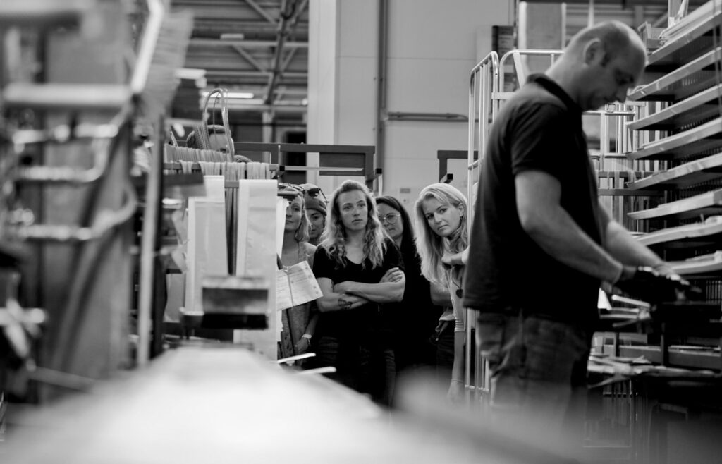 black and white photograph of a group of women watching a man tend to a stand of single skis on the production line in a factory setting