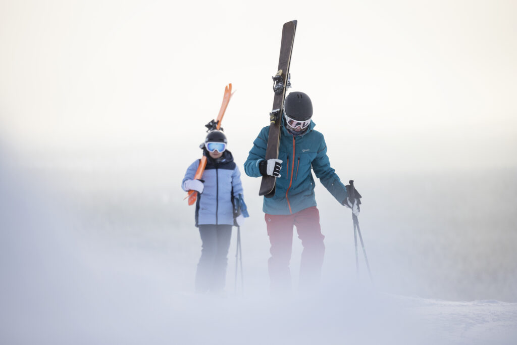 two skiers, with skis on shoulder walk through misty setting
