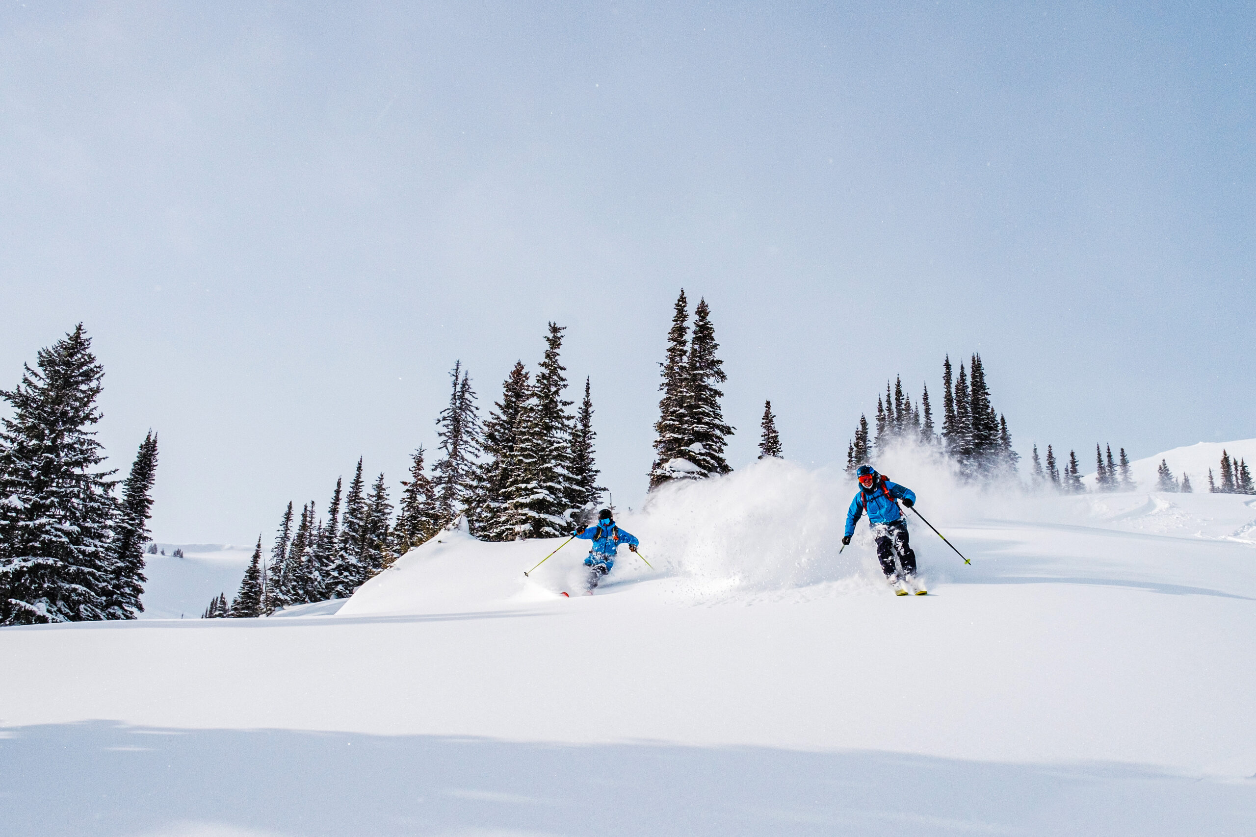 two skiers ski side by side on a mellow slope and untracked snow with mega powder spraying up behind skiers