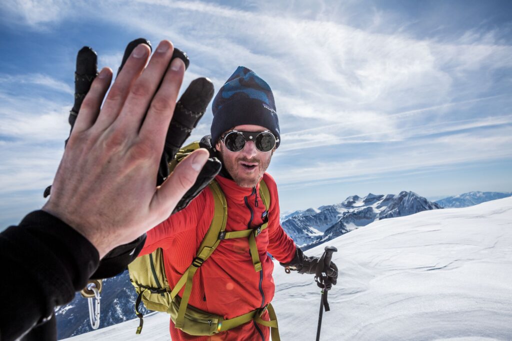 a man in vintage style alpine sunglasses gives a man with a bare hand (not in shot) a high-5 whilst ski touring