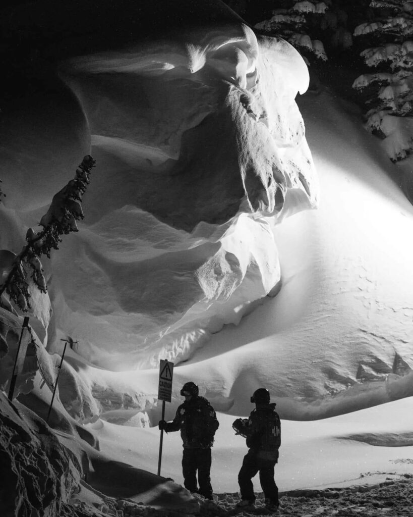 two patrollers are seen under the most enormous snow pile, in black and white image, snow rising metres upon metres above them like a monster