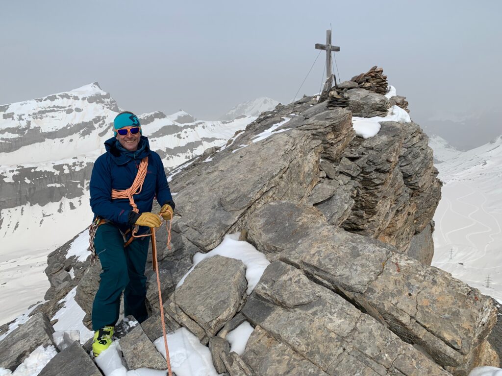 ski touring/mountaineer man stands at top of a mountain next to the cross that marks top on the rocky summit