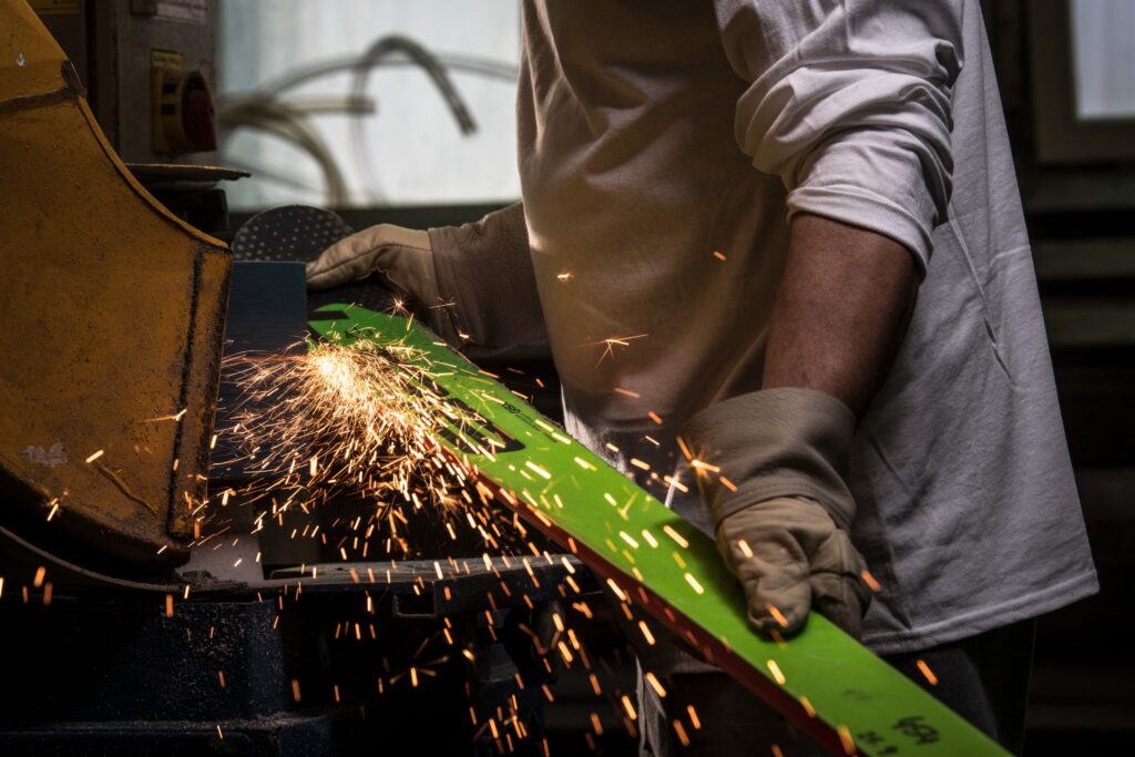 a green ski in production held by someone in gloves against a sanding machine of sorts, giving of sparks