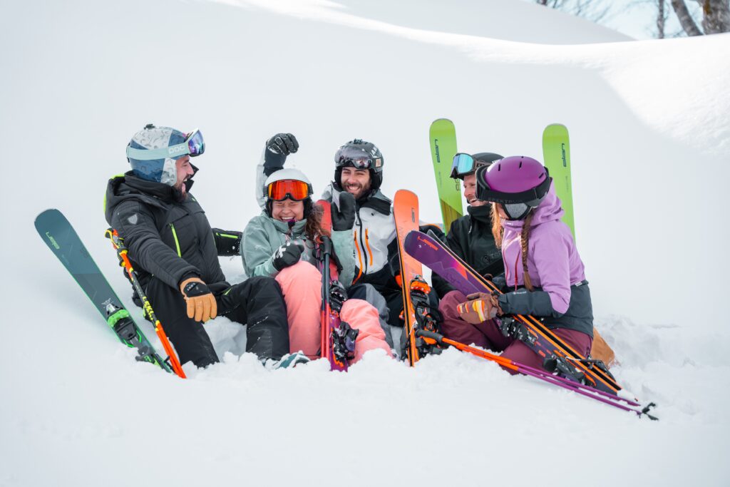 pile of skiers sit on snow dressed in colourful clothes all holding their Elan skis, laughing in a set up brand photo