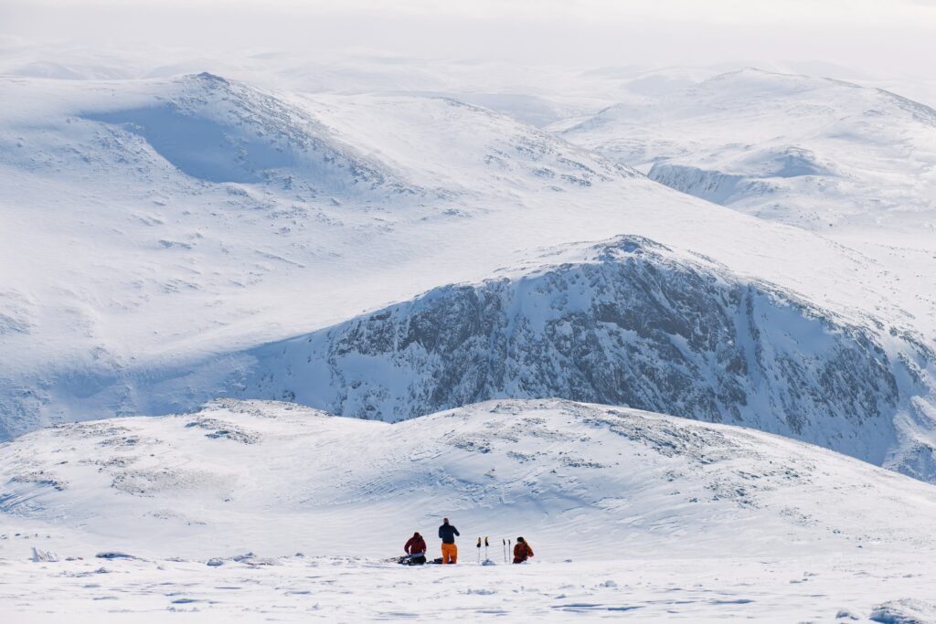 three ski tourers rest in the distance in front of the most mega view of snowy cairngorms, just three colourful dots on horizon, along with silhouette of poles